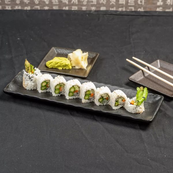 A black rectangular plate holds eight sushi rolls with asparagus, carrots, and rice, alongside wasabi, ginger, and chopsticks.