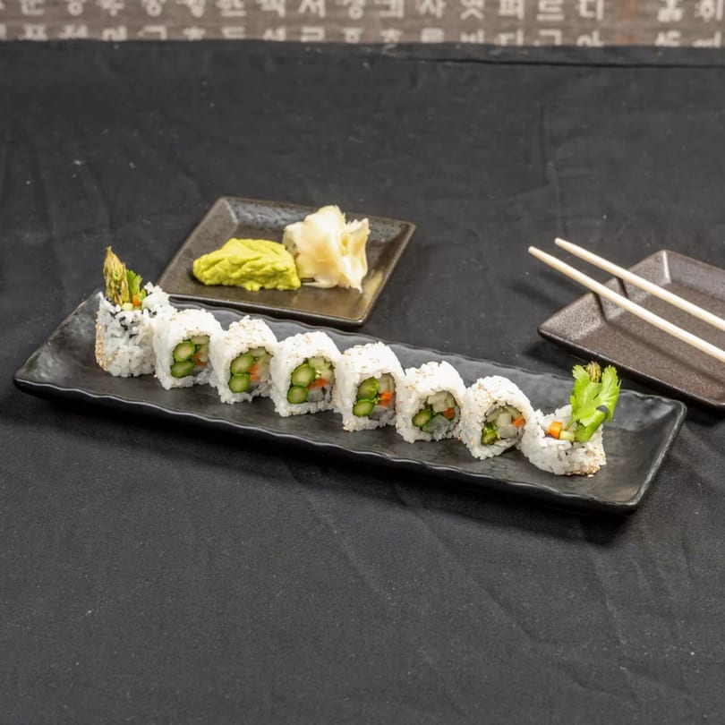 A black rectangular plate holds eight sushi rolls with asparagus, carrots, and rice, alongside wasabi, ginger, and chopsticks.