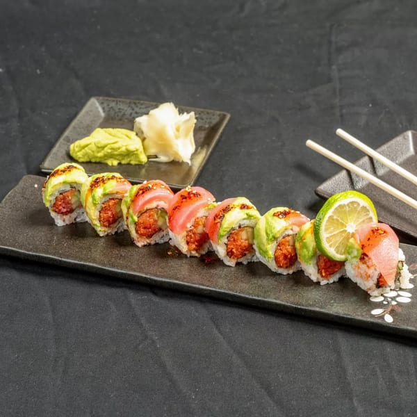 Sushi roll with avocado and tuna, served with wasabi, ginger, and chopsticks on a black plate.